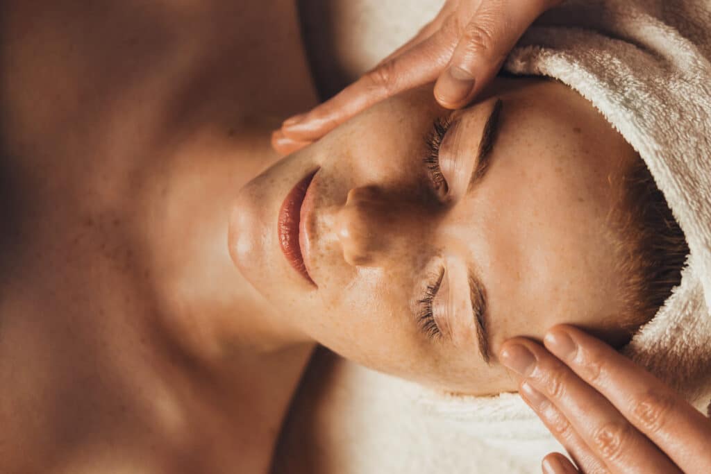 Massage du visage relaxant : détente et soin de la peau Femme rousse aux taches de rousseur recevant un massage du visage relaxant avec les yeux fermés, mains expertes sur son front.