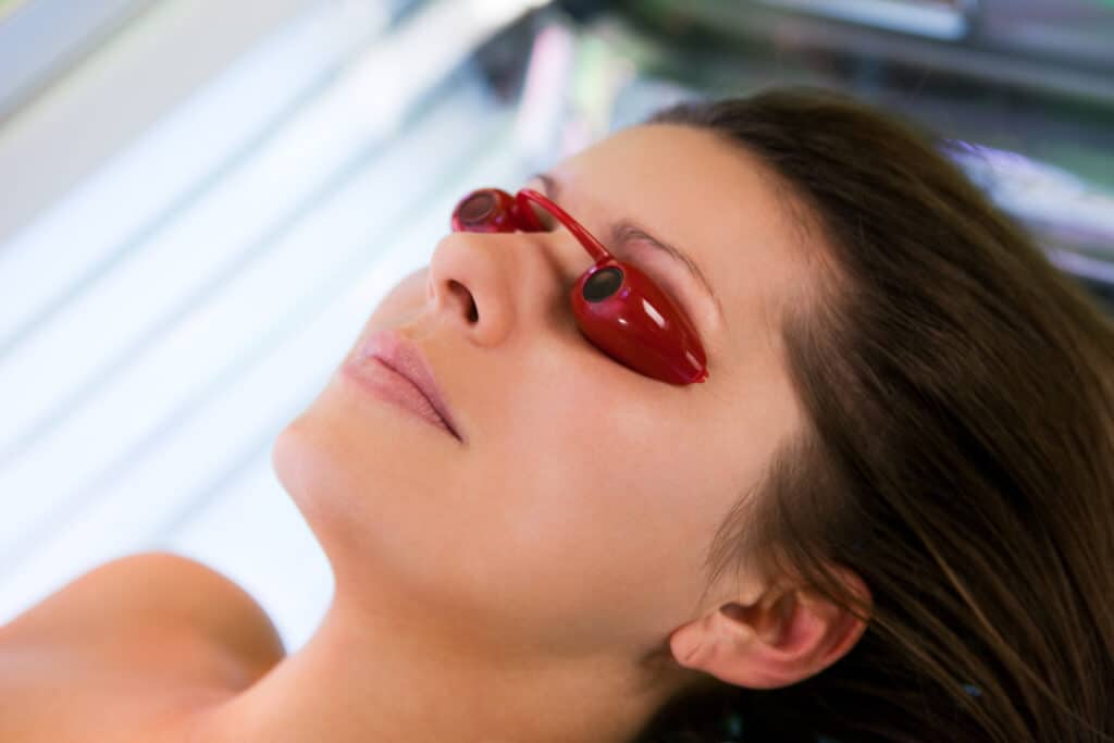 Bronzage UV: Femme avec lunettes de protection rouge Jeune femme au visage levé sous un bronzage, portant des lunettes de protection oculaires rouges dans un appareil de cabine de bronzage.